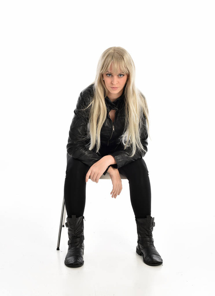Ganzkörperporträt eines blonden Mädchens in schwarzem Lederkostüm, sitzend. isoliert auf weißem Studiohintergrund. - Foto, Bild