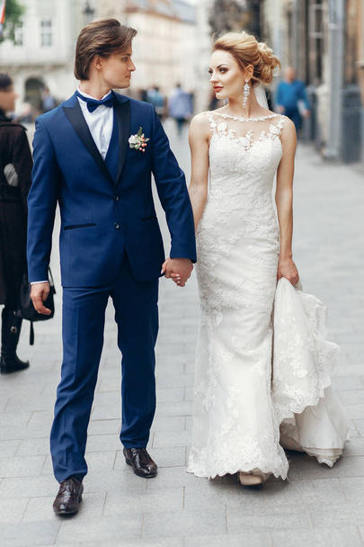 Κομψό νύφη και ο γαμπρός το περπάτημα στο δρόμο της πόλης. ευτυχισμένος πολυτελές γαμήλιο ζεύγος κρατώντας τα χέρια μέσα στο φως. Ρομαντικά αισθησιακό παρόν. άνδρας και γυναίκα βλέπουν ο ένας τον άλλον - Φωτογραφία, εικόνα