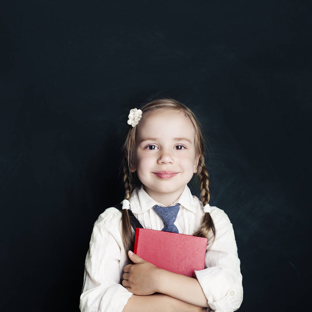 Happy little girl with red school book on a blackboard background.  Kid indoors of the class room. Back to school concep - Foto, Bild