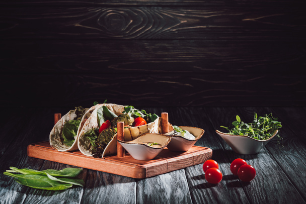 food composition of tortillas with falafel, cherry tomatoes and germinated seeds of sunflower on wooden tray with sauces on table - Photo, Image
