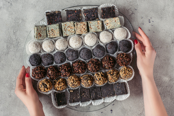 partial view of woman holding plate with various sweet desserts on grey tabletop - Foto, Imagem