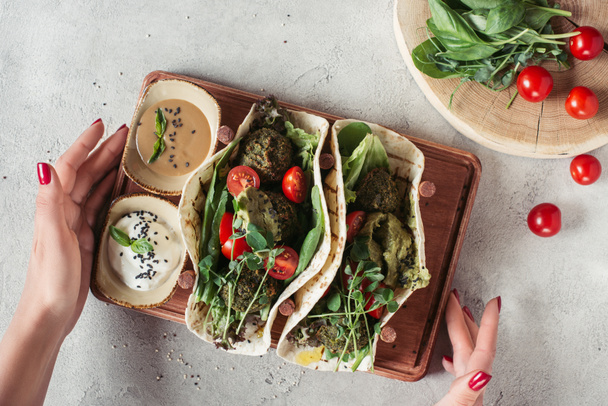 cropped shot of woman holding falafel with tortillas, cherry tomatoes and germinated seeds of sunflower served on wooden board on grey surface - Photo, Image