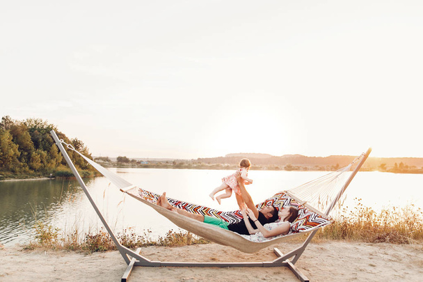 happy stylish family playing with cute daughter, relaxing in hammock on summer vacation in evening sun light on the beach. hipster couple with child resting and having fun in sunset. space for text - Fotoğraf, Görsel