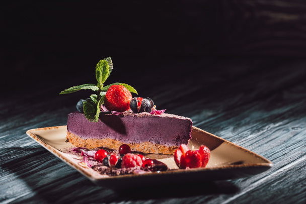 selective focus of blueberry cake with strawberries, mint and viola petals on plate on wooden table  - Foto, Imagen
