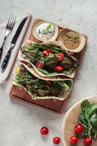 Draufsicht auf Falafel mit Tortillas, Kirschtomaten und gekeimten Sonnenblumenkernen, serviert auf Holzbrett auf grauer Oberfläche - Foto, Bild