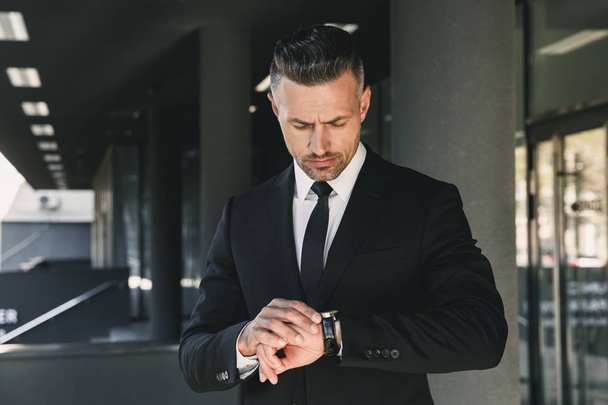 Portret uroczy młody biznesmen ubrany w garnitur, stojący na zewnątrz budynku ze szkła i patrząc na jego zegarek na rękę - Zdjęcie, obraz