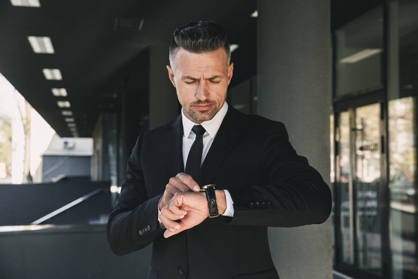 Portrait of a handsome young businessman dressed in suit standing outside a glass building and looking at his wrist watch - Φωτογραφία, εικόνα