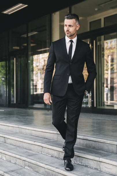 Portrait of a confident young businessman dressed in suit walking down the stairs outside a glass building - Fotografie, Obrázek