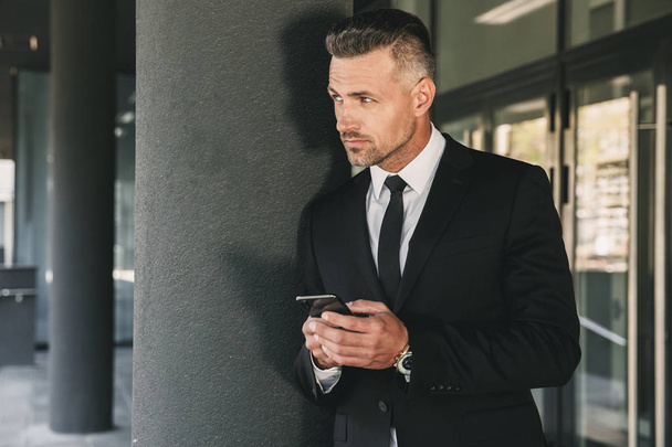 Portrait of a confident young businessman dressed in suit standing outside a glass building and using mobile phone - Photo, image