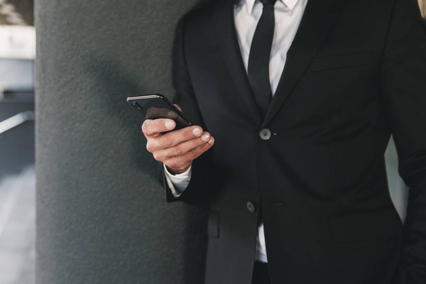 Imagen recortada de un hombre de negocios vestido de traje parado fuera de un edificio de vidrio y sosteniendo el teléfono móvil
 - Foto, imagen