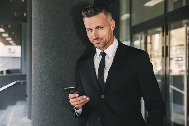 Porträt eines lächelnden jungen Geschäftsmannes im Anzug, der vor einem gläsernen Gebäude steht und Mobiltelefone benutzt - Foto, Bild