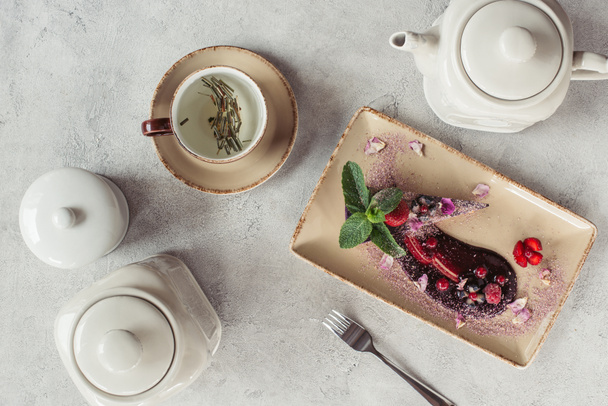 food composition with piece of blueberry cake served with mint leaves and violet petals on plate, teapot and cup of herbal tea on grey tabletop - Photo, Image