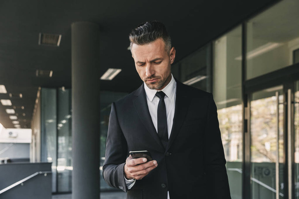 Portrait of a confident young businessman dressed in suit walking outside a glass building and using mobile phone - Фото, изображение