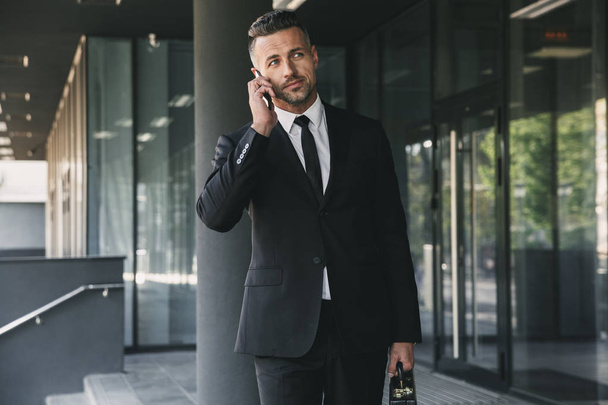 Portrait of a successful young businessman dressed in suit with briefcase walking outside a glass building and talking on mobile phone - Photo, Image