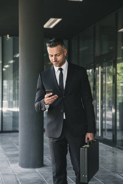 Portrait of a handsome young businessman dressed in suit with briefcase walking outside a glass building and using mobile phone - Photo, image