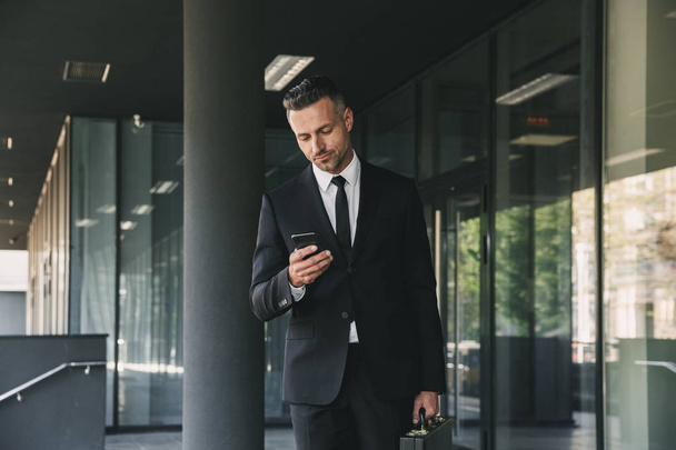 Portrait of a serious young businessman dressed in suit with briefcase walking outside a glass building and using mobile phone - Фото, изображение