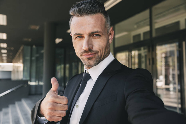 Portrait of a confident young businessman dressed in suit standing outside a glass building and taking a selfie - Φωτογραφία, εικόνα