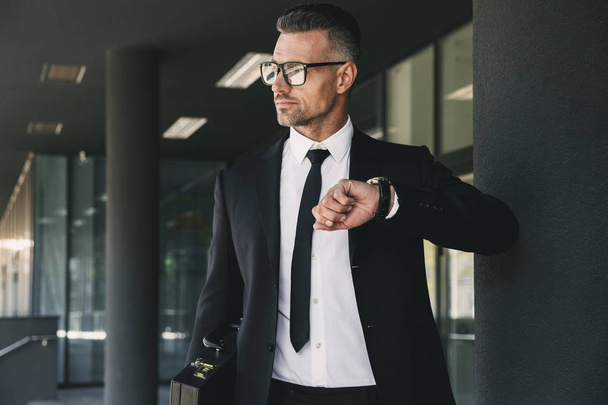 Portrait of a charming young businessman dressed in suit standing outside a glass building and looking at his wrist watch - Foto, imagen