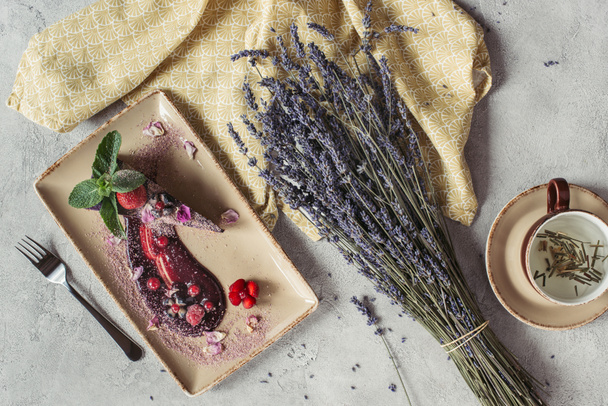 food composition with piece of blueberry cake served with mint leaves and violet petals on plate, lavender and cup of herbal tea on grey tabletop - Photo, Image