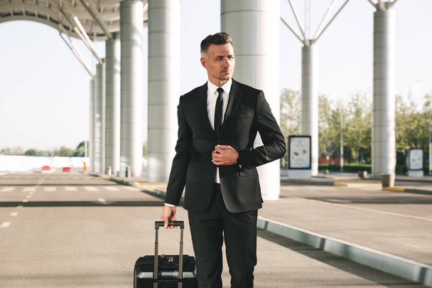Confident businessman dressed in suit walking with a suitcase outside airport terminal - Photo, image