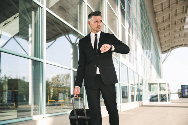 Relaxed businessman dressed in suit walking with a suitcase outside airport terminal and looking at his wristwatch - Foto, Imagem