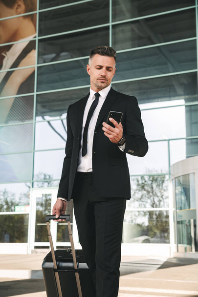 Handsome businessman dressed in suit walking with a suitcase outside airport terminal and holding mobile phone - Foto, imagen