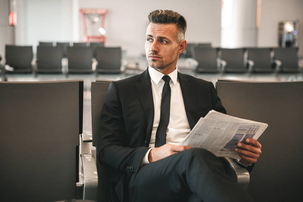 Seriöser Geschäftsmann liest Zeitung, während er in der Lobby des Flughafenterminals sitzt - Foto, Bild