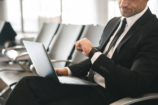 Cropped image of businessman dressed in suit using laptop computer and checking time while sitting at the airport lobby - Foto, afbeelding