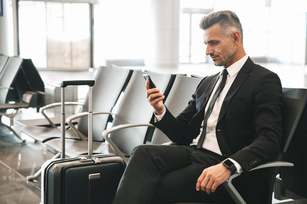 Handsome mature businessman using mobile phone while sitting at the airport lobby with a suitcase - Fotografie, Obrázek