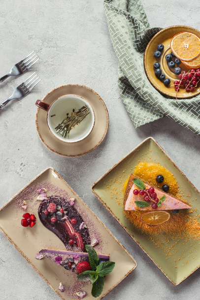 platte lay met zoete worteltaart met bessen vullen, bosbessen cake geserveerd met muntblaadjes en violet bloemblaadjes, kopje kruidenthee en bestek op grijs tafelblad - Foto, afbeelding