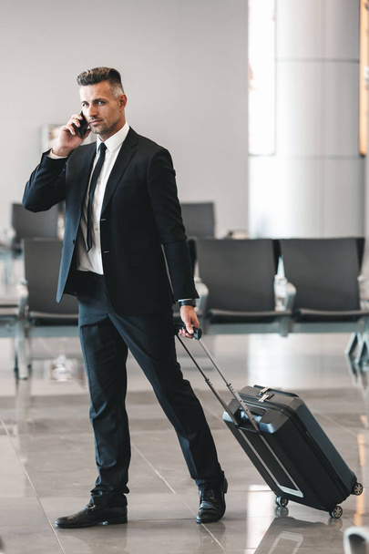 Handsome mature businessman talking on mobile phone while walking at the airport lobby with a suitcase - Fotoğraf, Görsel