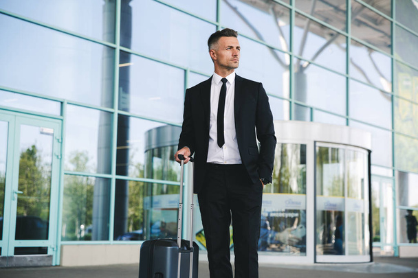 Serious mature businessman standing outside the airport with a suitcase - Photo, image