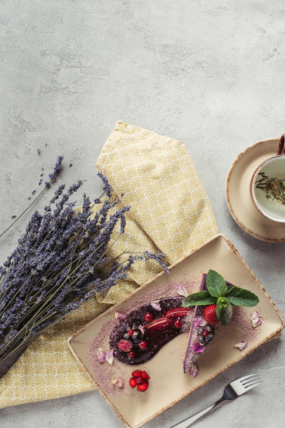 food composition with piece of blueberry cake served with mint leaves and violet petals on plate, lavender and cup of herbal tea on grey tabletop - Photo, Image