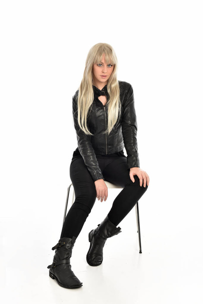 Ολόσωμο πορτρέτο του ένα ξανθό κορίτσι φορώντας μαύρα Δερμάτινα ρούχα, καθιστή στάση. απομονωμένα σε φόντο λευκό στούντιο. - Φωτογραφία, εικόνα