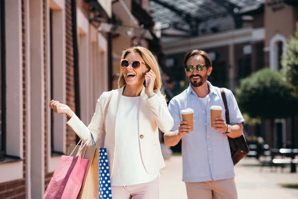 beszélő smartphone and gazdaság bevásárló táskák, férfi és eldobható kávéscsészét utcán boldog nő - Fotó, kép