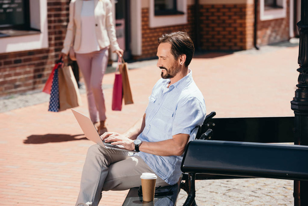 handsome man sitting on bench with laptop, woman walking with shopping bags on street - Photo, Image