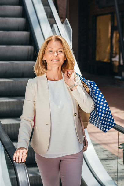 Alışveriş merkezinde alışveriş torbaları ile yürüyen merdiven üzerinde duran gülümseyen çekici kadın - Fotoğraf, Görsel
