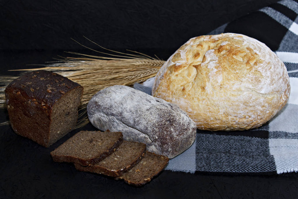 Sortidas diferentes tipos de pão branco e preto em um fundo preto ao lado das orelhas de trigo
. - Foto, Imagem