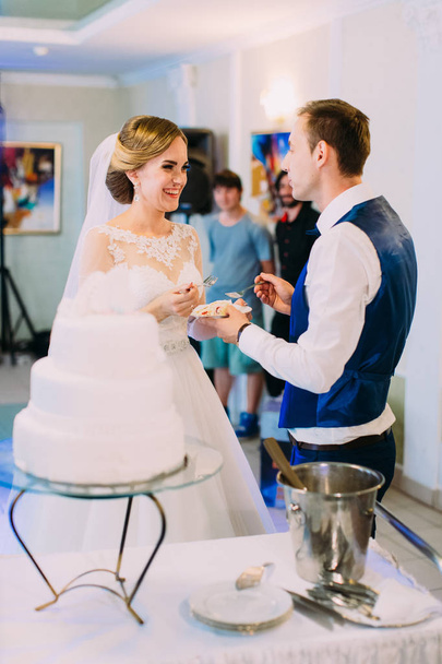 Smiling newlywed couple is feeding each other with the wedding cake. - Фото, изображение