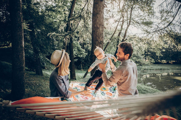 家族の幸せな瞬間。夏休み。流行に敏感な家族が夏の日当たりの良い公園のハンモックにはかわいい息子と遊ぶ。スタイリッシュなママ ・ パパ、そして子供のリラックス。テキストのためのスペース. - 写真・画像