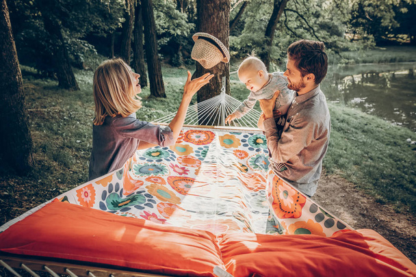 happy hipster family playing with cute little son on hammock in summer sunny park. stylish mom and dad with kid relaxing in forest and smiling, having fun. space for text. family vacation - Photo, Image