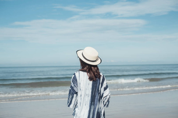 旅行者のビーチで一人帽子立っているとカジュアルな服装の若い女性が青い空と海の背景 - 写真・画像