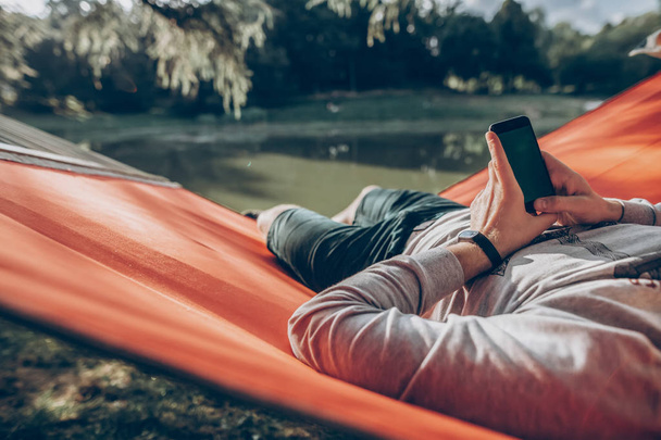 hipster man holding smart phone with empty screen with space for text, and relaxing in hammock in sunny summer park. mock up. guy looking at blank phone, technology communication concept - Photo, image
