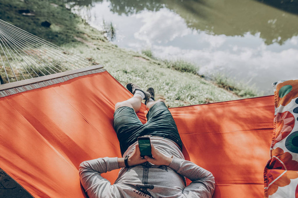 hipster man holding smart phone with empty screen with space for text, and relaxing in hammock in sunny summer park. mock up. guy looking at blank phone, technology communication concept - Photo, Image