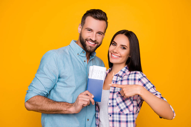 Portrait of joyful glad couple holding passport with flying tickets in hands pointing with forefinger looking at camera isolated on bright yellow background - Foto, immagini