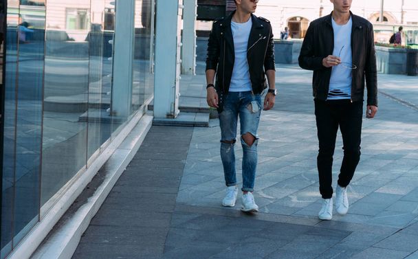 beautiful and attractive guy is going in the city, posing. dressed in a black leather kurta, white T-shirt, jeans and white shoes. near the mirror building. walk around the city in black glasses. communication. meeting friends. - Foto, Imagen