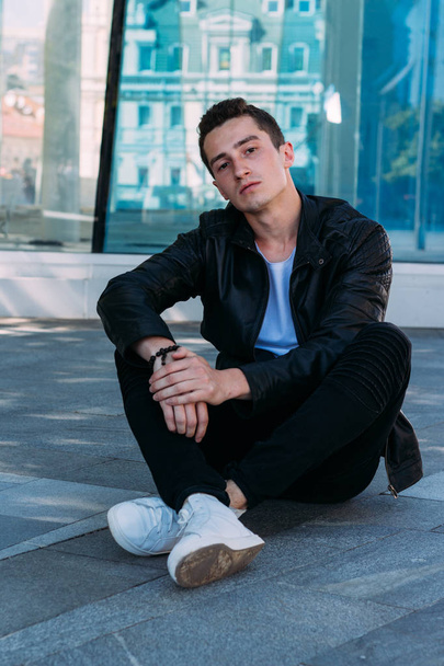 beautiful and attractive guy is sits near a glass building on the floor in the city, posing. dressed in a black leather kurta, white T-shirt, jeans and white shoes. city Style - Φωτογραφία, εικόνα