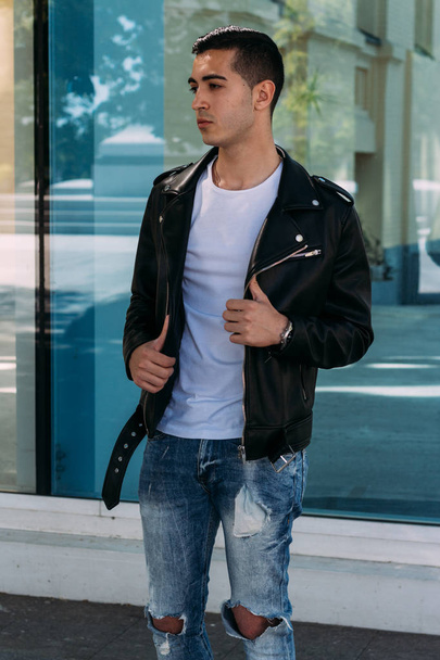 beautiful and attractive guy is standing near the glass building in the city, posing. dressed in a black leather kurta, white T-shirt, jeans and white shoes. - Fotografie, Obrázek