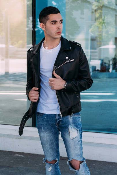 beautiful and attractive guy is standing near the glass building in the city, posing. dressed in a black leather kurta, white T-shirt, jeans and white shoes. - Foto, imagen
