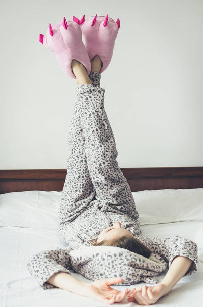 Young woman enjoying sunny morning. Caucasian girl in cute warm pajamas and pink monster foot slippers with big claws lying on the bed at home bedroom after waking up. Good start of the day concept. - Photo, Image